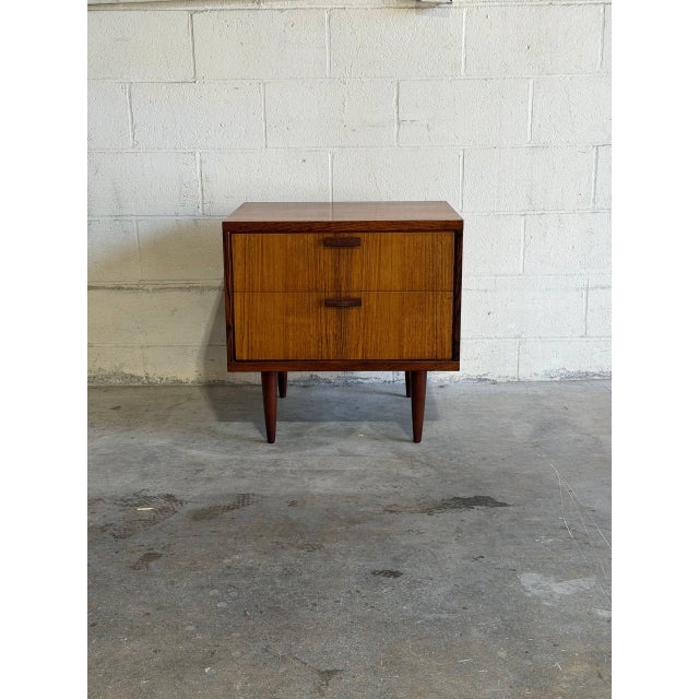 This architectural bedside chest features a compact box frame elevated by slender tapered legs. Two smooth pull-out...