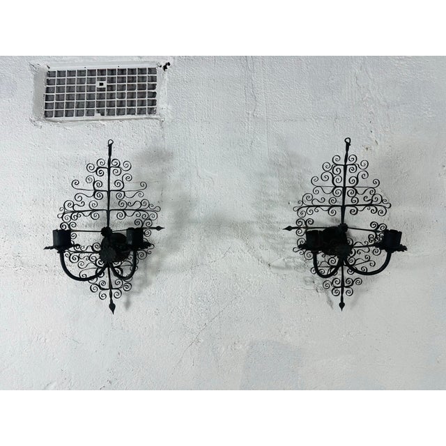 A striking pair of vintage hand-forged wrought iron two-light wall sconces featuring intricate scrolling filigree...