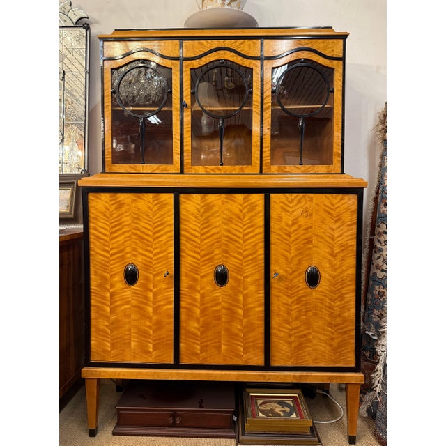 This tall vitrine sideboard combines a warm, highly figured birch veneer with ebonised mouldings and striking circular...