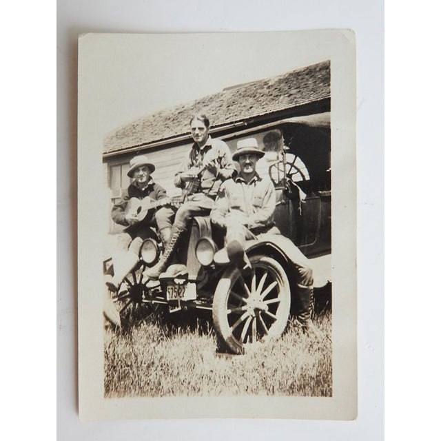 Early 20th century sepia snapshot photograph of three men sitting on the hood of a car playing a guitar and banjo....