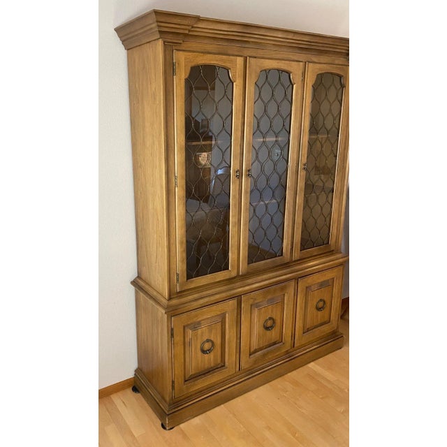A mid-century Drexel china cabinet in a warm honey-brown wood finish. The upper hutch has three glazed doors with arched...
