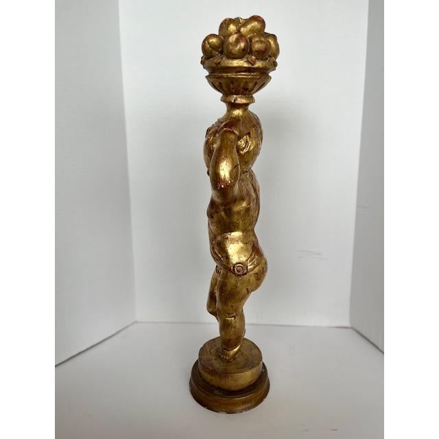 wooden gold statue of a putti holding a bowl of fruits on top of his head. the wood seeps through the gold paint giving it...