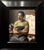 The painting depicts a contemplative figure seated in an artist's studio, holding a cup while gazing thoughtfully at a...