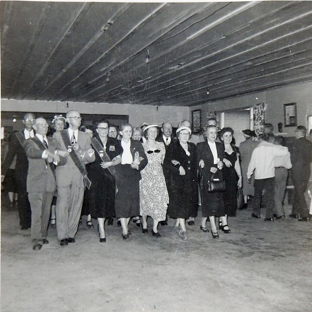 Mid 20th century snapshot photograph of group of people line dancing in a large hall. Ink note says Rural Life Sunday....