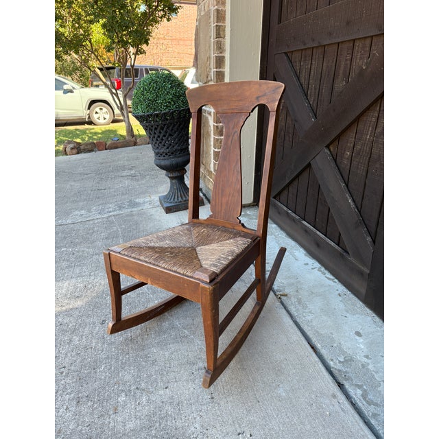 This charming early 20th-century wooden rocking chair features a beautiful Arts & Crafts design with a handwoven rush seat...