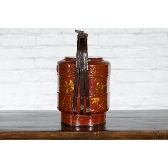 A Chinese three-tiered lunch basket from the early 20th century with red lacquer, gilded décor and black bamboo handle....