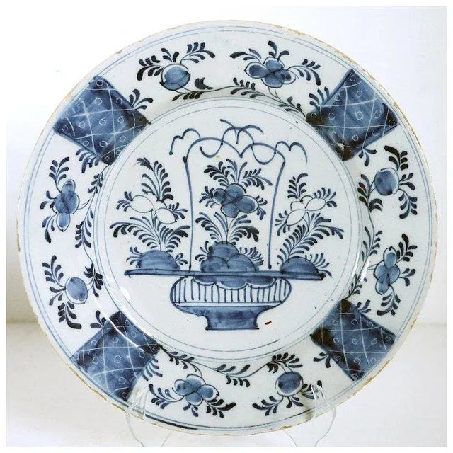 DETAILS An antique Dutch Delft faience plate with an elaborate floral basket arrangement in the center. It is bordered by...
