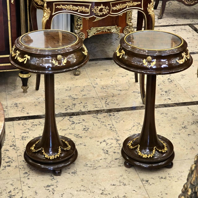 A sophisticated matching pair of French Neoclassical side tables, featuring rich dark wood finishes, glass-inset tops, and...