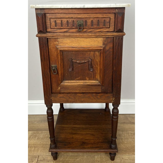 A charming antique French bedside table featuring a beautiful white marble top above a richly grained oak body with carved...