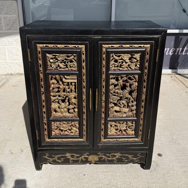 This heavily carved black Asian cabinet features intricately detailed gold doors with relief scenes framed by ornate...