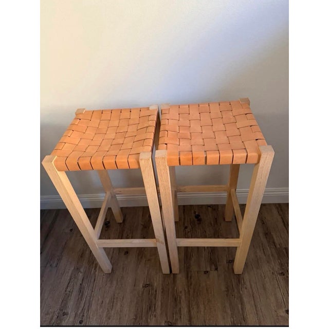 A beautifully pair of bar stools featuring thick, handwoven natural tan leather seats over solid oak frames in a light...