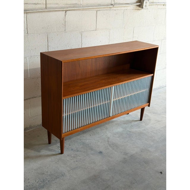Beautiful vintage Danish modern teak cabinet featuring sleek tambour sliding doors, open shelving, and elegant tapered...