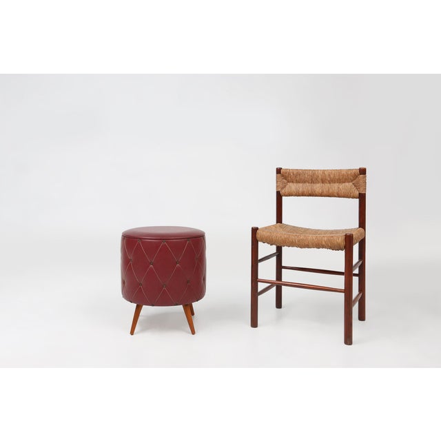 Vintage pouf made of dark red artificial leather with a padded look. At the bottom, the ottoman rests on four wooden legs....