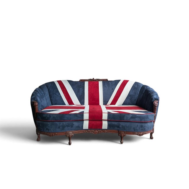 A striking upholstered sofa pairing a bold Union Jack motif in blue, red, and cream with an ornate carved wood frame. The...