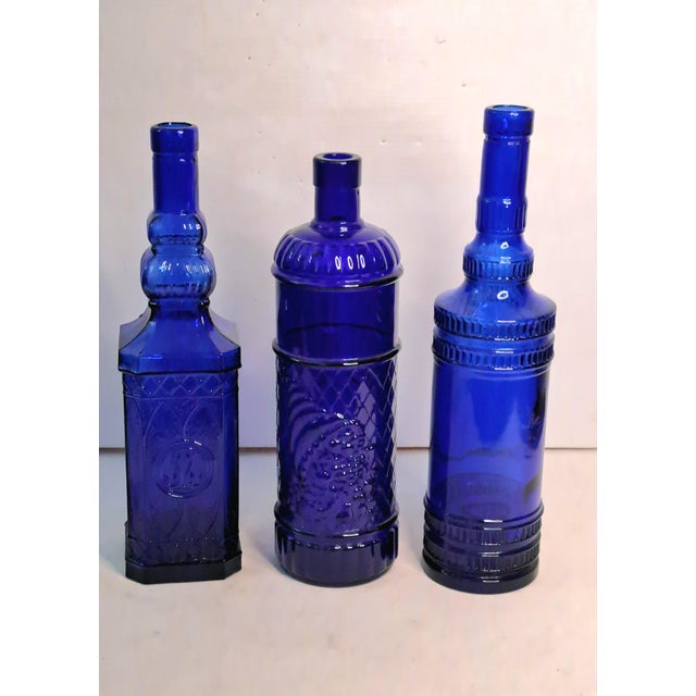 A striking collection of three vintage cobalt blue art glass bottles featuring distinct architectural forms, embossed...