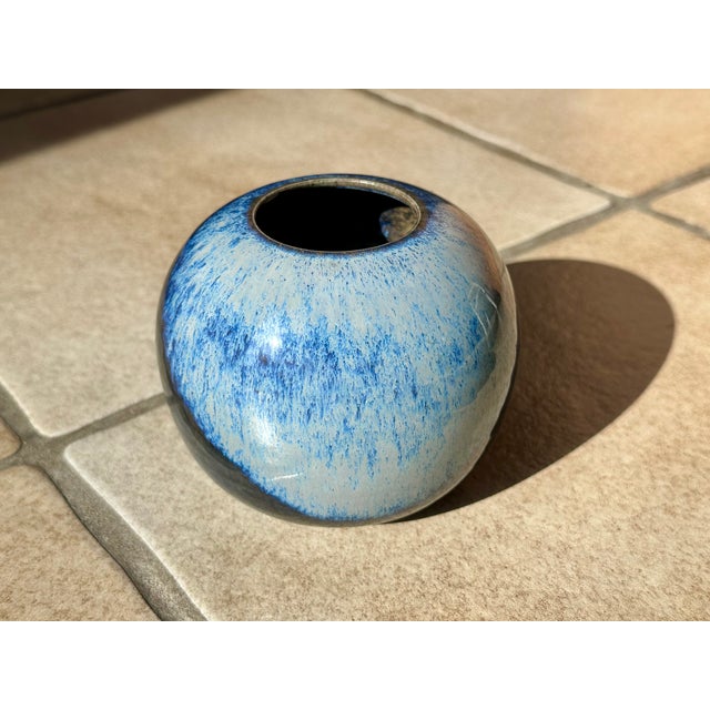 Handcrafted ceramic spherical vase with a striking blue-gray flowing glaze and a glossy finish on a light-colored body....
