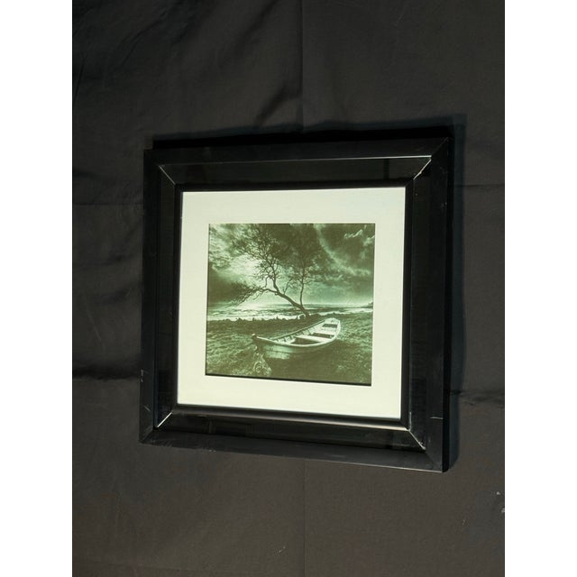 Elegant black and white framed landscape photograph featuring a small boat, tree, shoreline, and dramatic outdoor scenery....