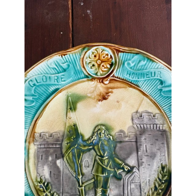 French majolica commemorative plate with relief-molded center depicting Joan of Arc before a fortified tower, holding her...