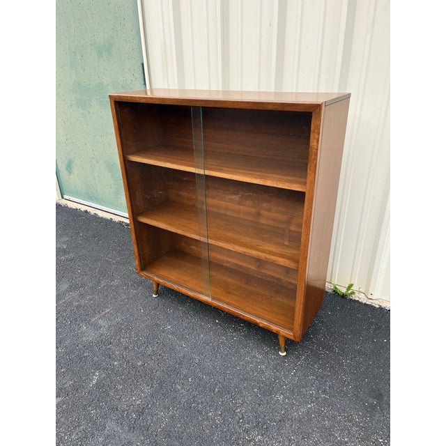 A vintage walnut double door bookcase with sliding glass doors. Features two shelves for a total of three interior storage...