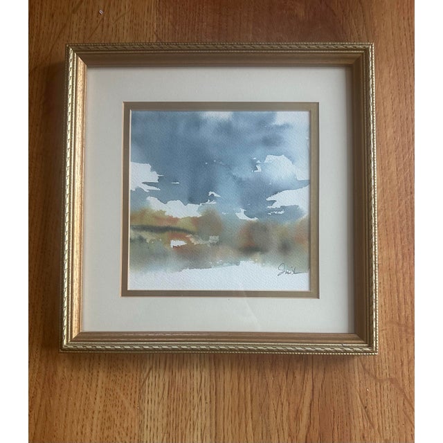 Soft washes of blue and muted earth tones suggest a calm horizon beneath drifting clouds. This watercolor captures a quiet...