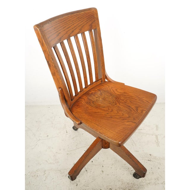 Antique swivel quarter sawn oak office chair with a dark stain. It features a vertical back slatted design, a contoured...