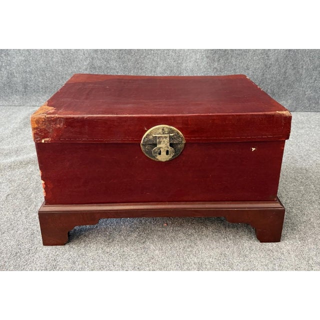 This vintage trunk is wrapped in worn red leather with a brass clasp and side handles, resting on a fitted wooden stand....