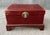 This vintage trunk is wrapped in worn red leather with a brass clasp and side handles, resting on a fitted wooden stand....