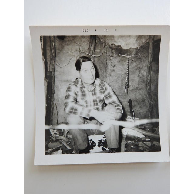 1970 snapshot photograph of man inside a Native American shelter. He is wearing a plaid western shirt, the shelter looks...