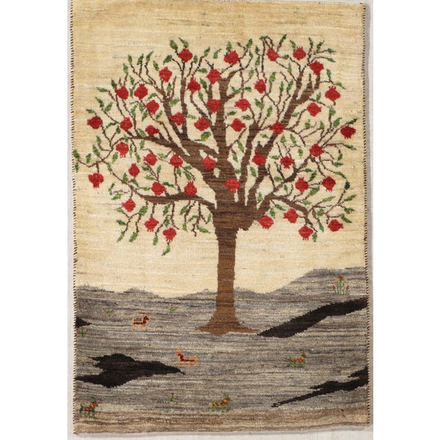 Hand-knotted Persian Gabbeh wool rug depicting a large tree with red fruit on an ivory field, set above a landscape with...