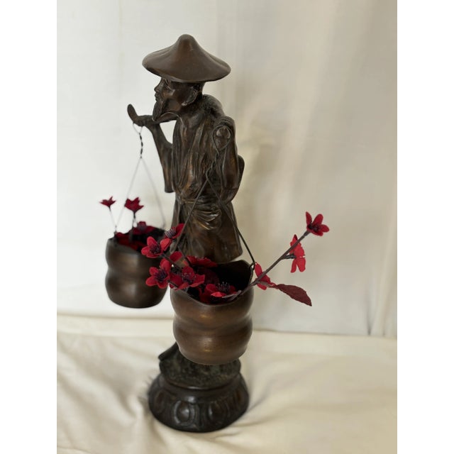 Bronze statue of elderly man wearing a traditional hat and flowing robe, carrying two buckets suspended from yoke across...