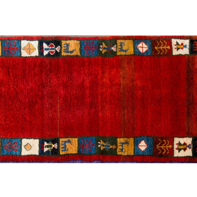 A gorgeous mid-20th century Persian Gabbeh rug with a bold abrash crimson field surrounded by myriad squares with stylized...