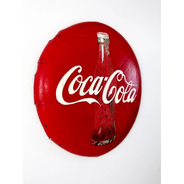 This vintage Coca-Cola advertising sign features a large, three-dimensional metal button design showcasing the iconic red...