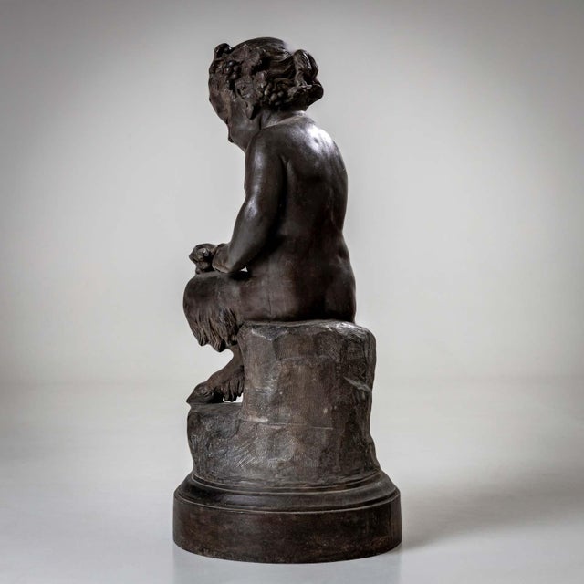 Black sculpture of a young satyr on a pedestal stroking a lizard. His forehead is adorned with horns, his head with a...