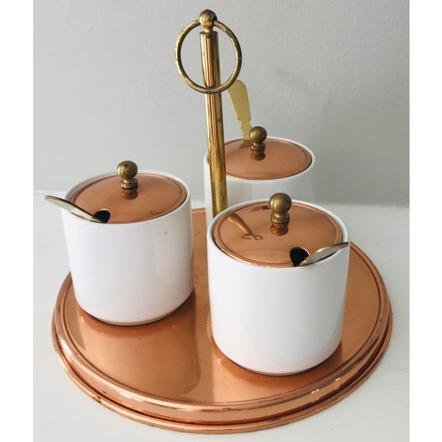 This is a vintage condiment server with three white ceramic cups, brass spoons and handle, copper lids, and a copper...
