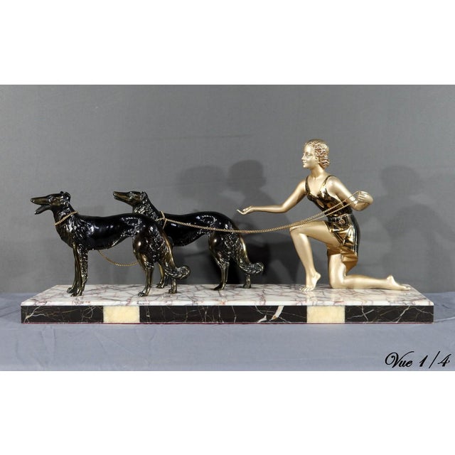 This work represents a woman accompanied by two greyhounds, made of metal with a patinated finish, black and gold in...