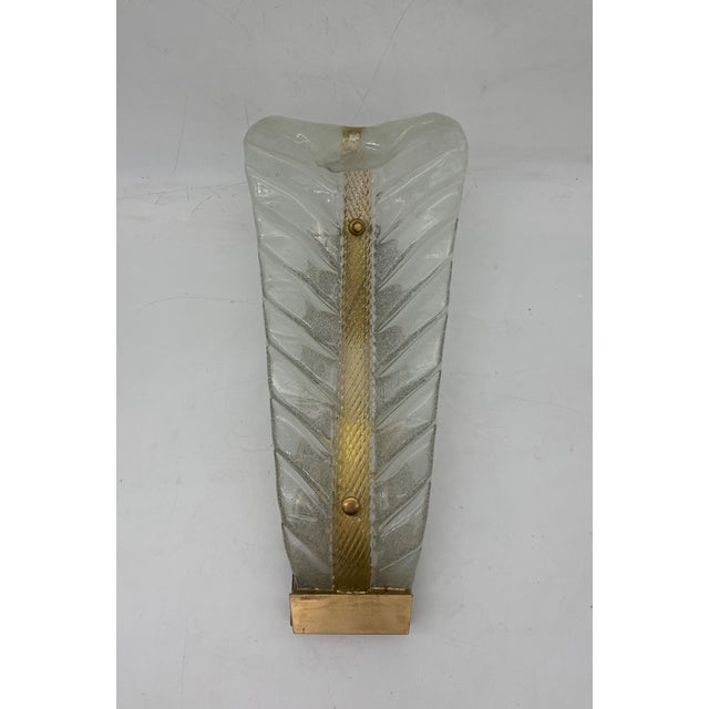 The lamp is a decorative wall light with a sculptural design inspired by the shape of a stylized leaf. Crafted from clear...