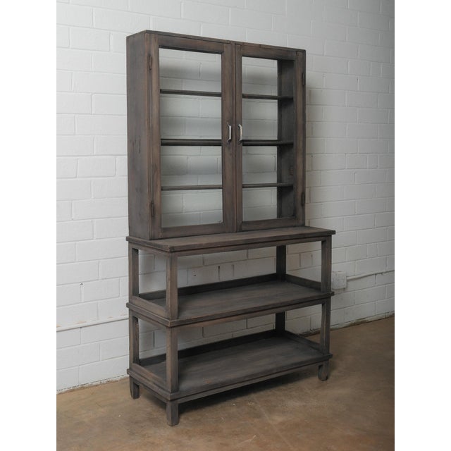 A quietly architectural glass-front cabinet set above a tiered open base, crafted in a deep, charcoal-toned wood that...