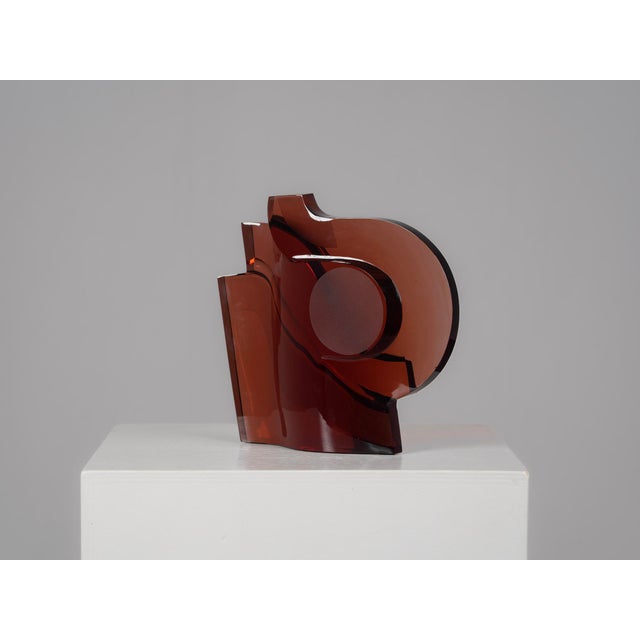 A striking abstract glass sculpture composed of strong geometric forms and flowing curves. The deep red-brown transparent...