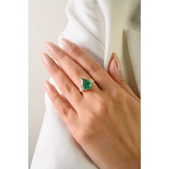 This elegant three-stone engagement ring features one natural pear-cut emerald weighing approximately 3.47 carats,...