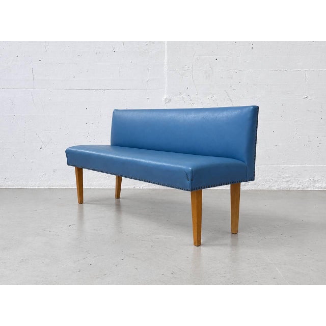 A characterful vintage bench with a blue faux leather cover, a straight backrest, and slender wooden legs. The minimalist...
