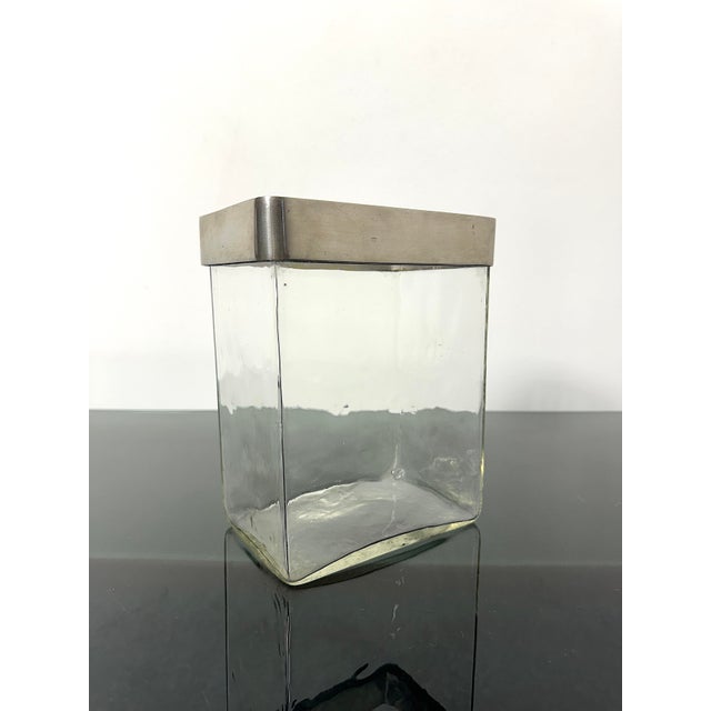 Flower vase composed of a transparent glass container with a rectangular base and a decorative upper edge in satin metal....