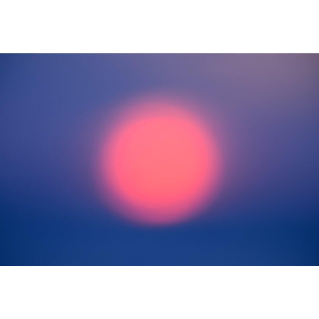 This photograph captures a soft, ethereal sunset, where the sun appears as a glowing, pink orb against a gradient of deep...