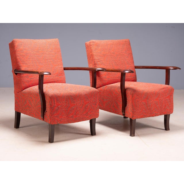 A distinctive pair of vintage mid-century armchairs with sculpted wooden armrests, tapered wooden legs, and red-orange...