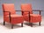 A distinctive pair of vintage mid-century armchairs with sculpted wooden armrests, tapered wooden legs, and red-orange...