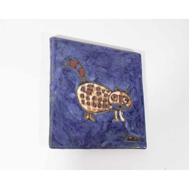 A Mexican ceramic tile trivet of a cat. The trivet depicts a spotted brown and white cat standing against a blue...