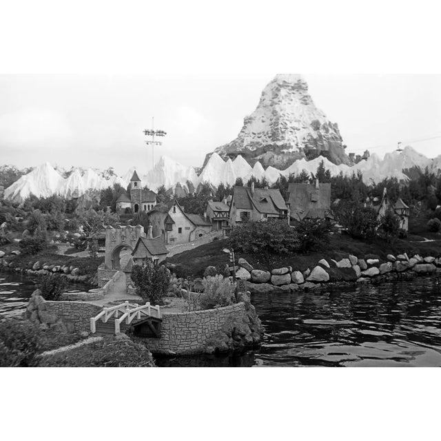 This black-and-white photograph captures the whimsical beauty of Disneyland’s Storybook Land with the towering Matterhorn...