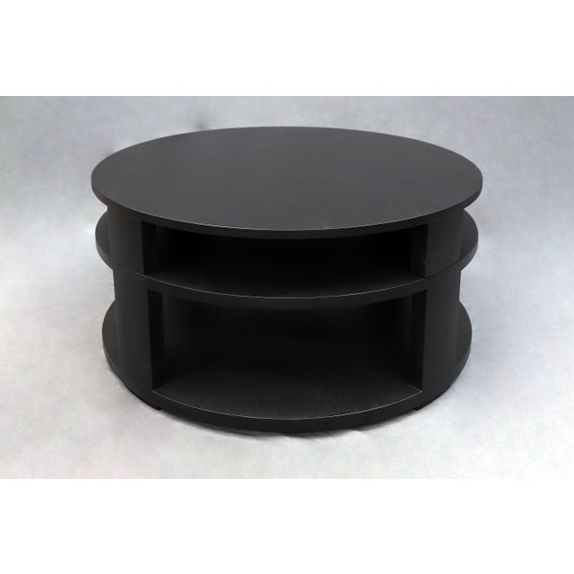 Circular wooden coffee table with black color finish. Middle shelf and a bottom shelf low to the ground.