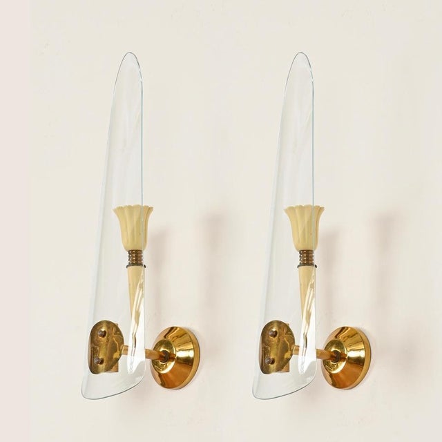 Elegant and refined pair of mid-century italian wall sconces in brass, cream enameled metal and curved glass. This very...