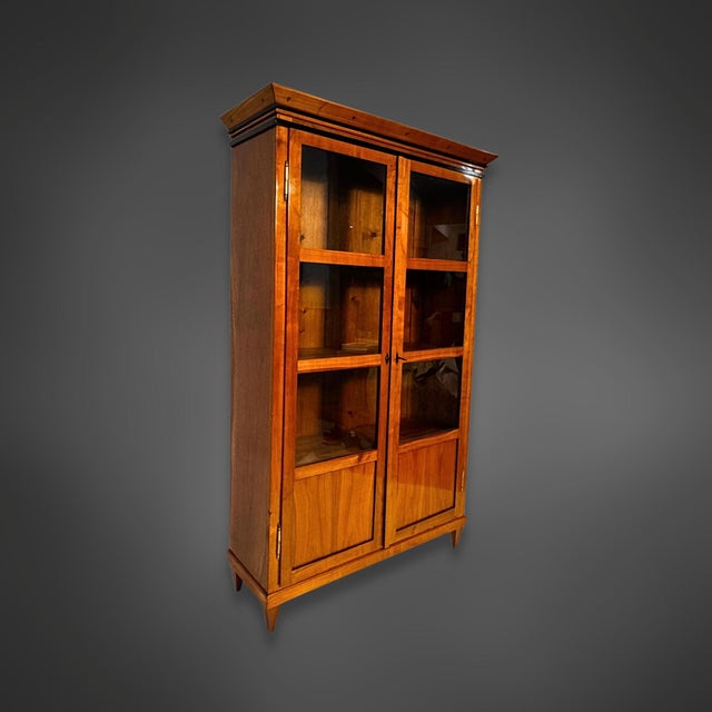 A two-door Biedermeier display cabinet in cherrywood. The cabinet's design is simple, crowned by a projecting cornice with...