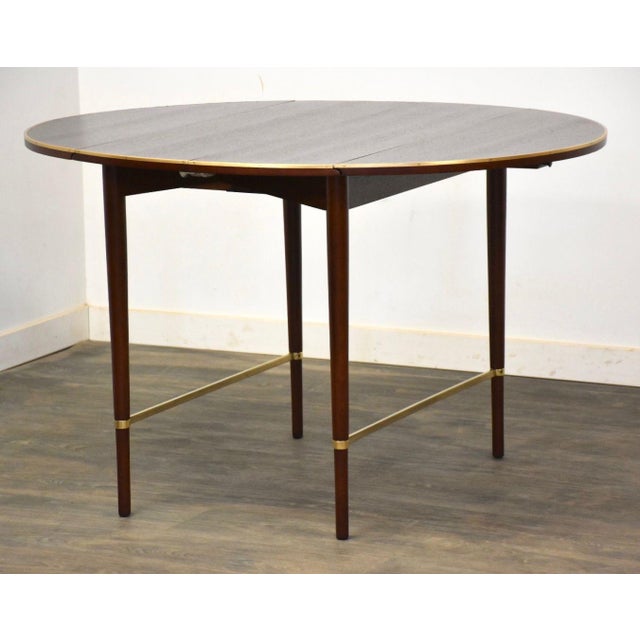 A mid century modern mahogany and brass oval “Connoisseur” drop leaf dining table designed by Paul McCobb for Calvin....